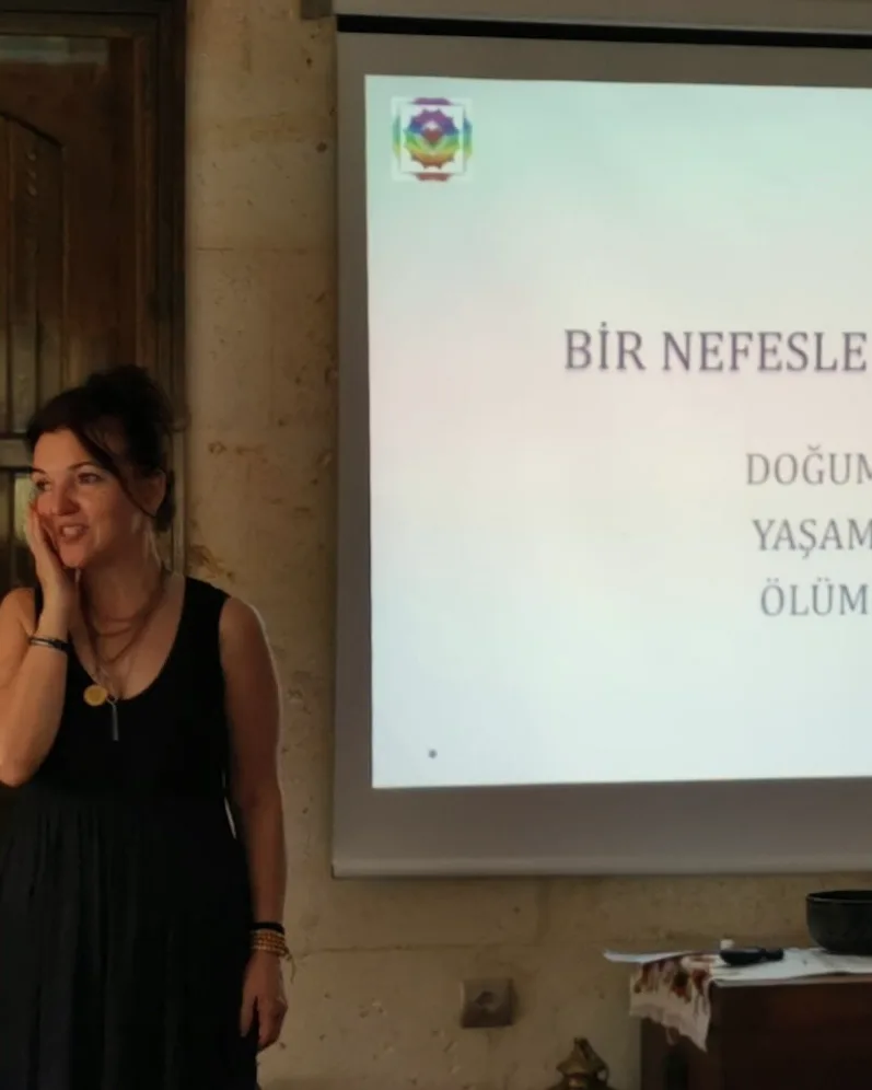 Arkasındaki perdede “Bir nefesle başladı: doğum, yaşam, ölüm” yazan sunumun önünde gülümseyen bir eğitmen